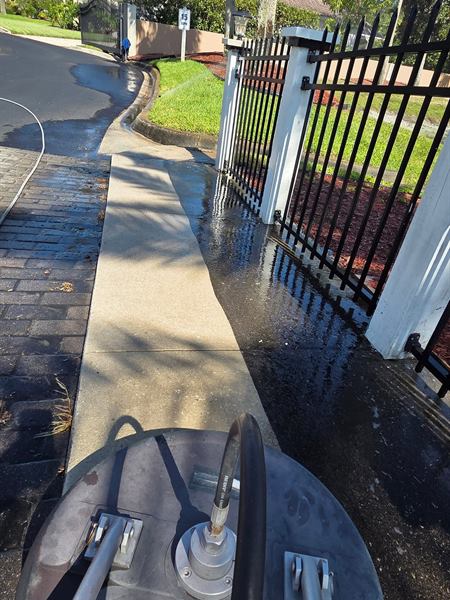 A surface cleaner partway along a community sidewalk beside an iron fence, the cleaned section brighter